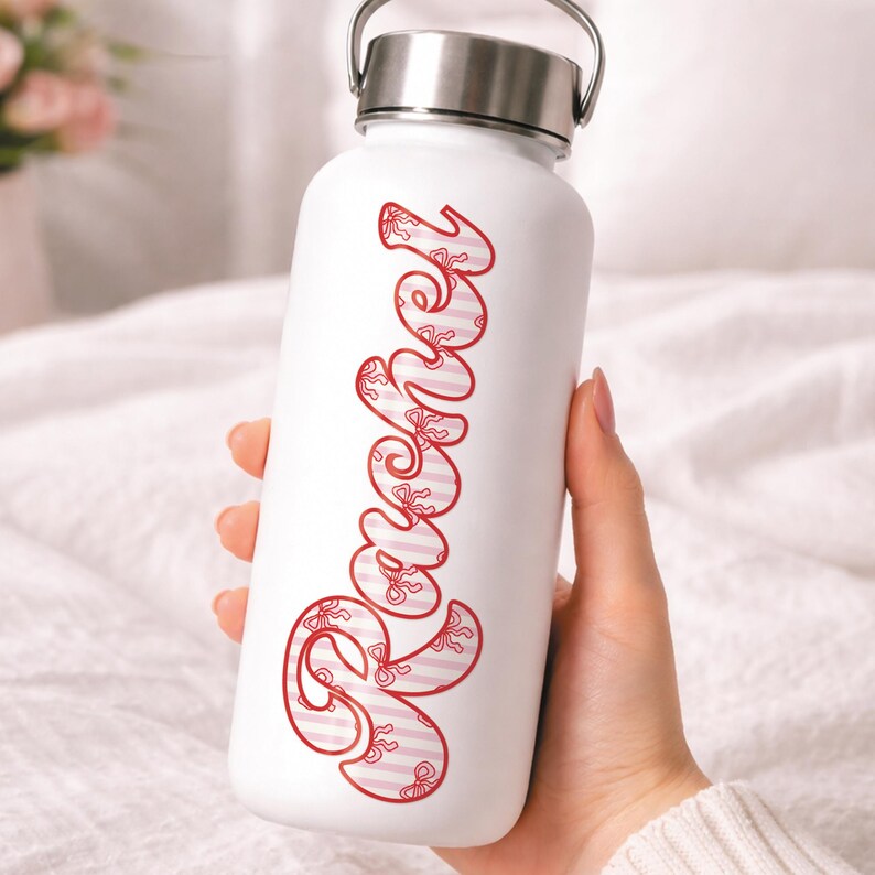 May include: White water bottle with a silver lid and handle. The word "Sweet" is written vertically in red script with a striped and bow pattern. The bottle is held by a person.