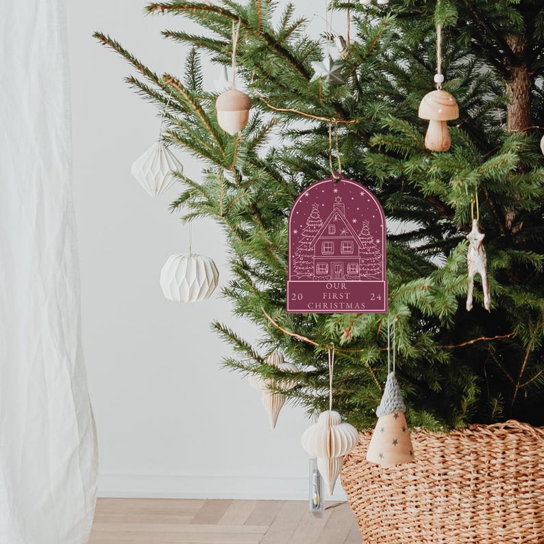 May include: Burgundy red wooden Christmas ornament with a white outline of a house and the text "Our First Christmas 20 24".