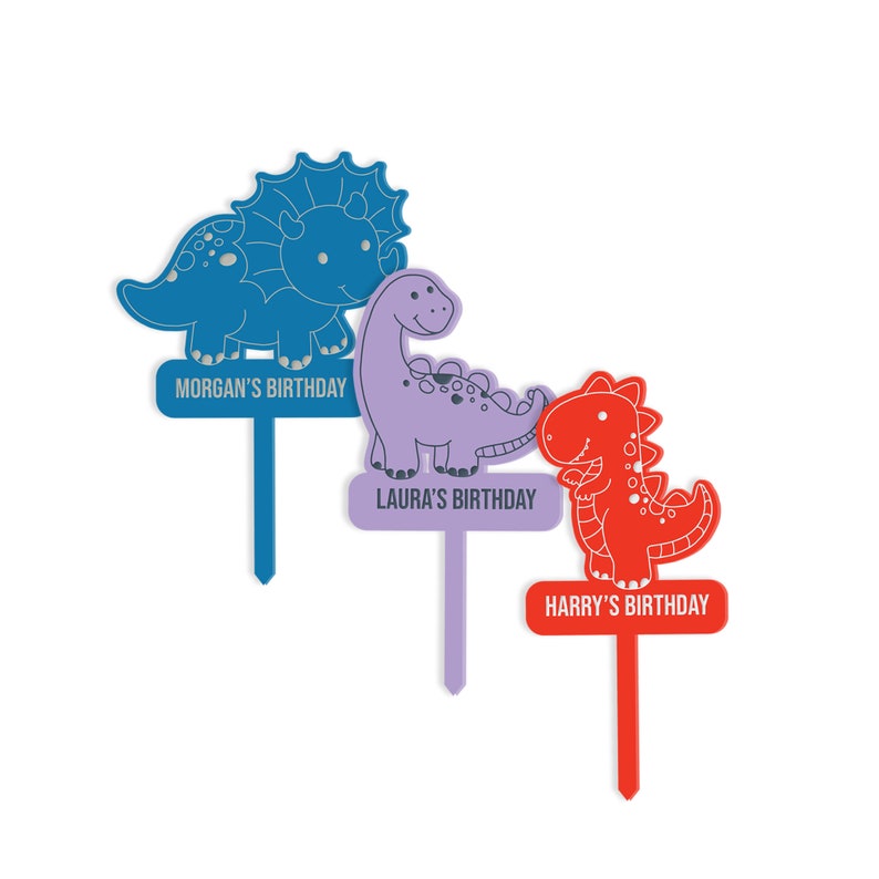 May include: Three colorful dinosaur cake toppers with personalized birthday messages. The blue dinosaur is a triceratops, the purple dinosaur is a brachiosaurus, and the red dinosaur is a tyrannosaurus rex.