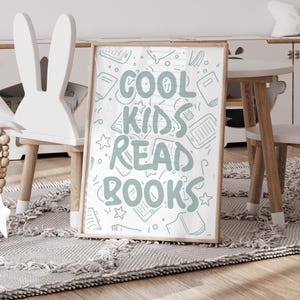 May include: A framed print with the words "COOL KIDS READ BOOKS" in light blue, surrounded by book illustrations. The print is in a wooden frame. The image is set in a child's room, with a white bunny chair and a small table.