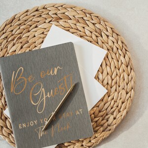 Pu&ograve; includere: Un diario grigio con lettere dorate che recitano "Be our Guest" e "Enjoy your stay at The Nook". Una penna dorata &egrave; appoggiata sul diario. Il diario &egrave; su un sottopentola di paglia intrecciata.