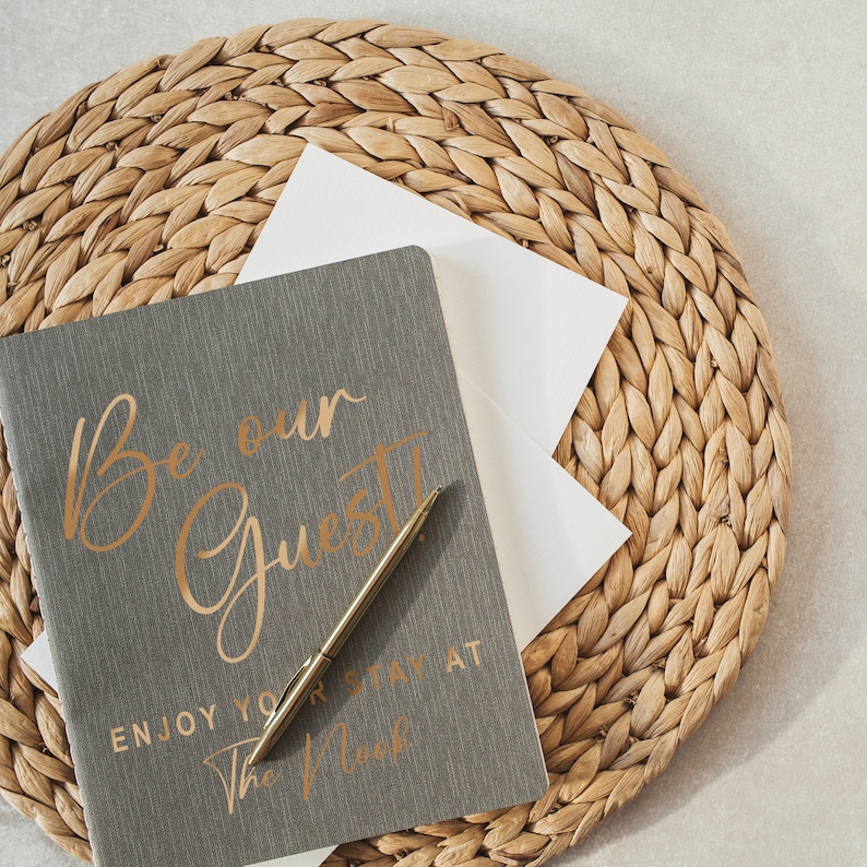 Pu&ograve; includere: Un diario grigio con lettere dorate che recitano "Be our Guest" e "Enjoy your stay at The Nook". Una penna dorata &egrave; appoggiata sul diario. Il diario &egrave; su un sottopentola di paglia intrecciata.