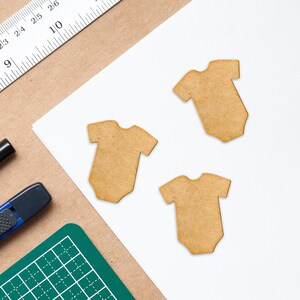May include: Three brown wooden baby onesie shapes on a white background. The onesies are cut out and have a simple design.