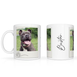 May include: White ceramic mug featuring a photo of a gray dog with its tongue out, wearing a pink collar. The mug has the name "Buster" in cursive on the side and a paw print design.