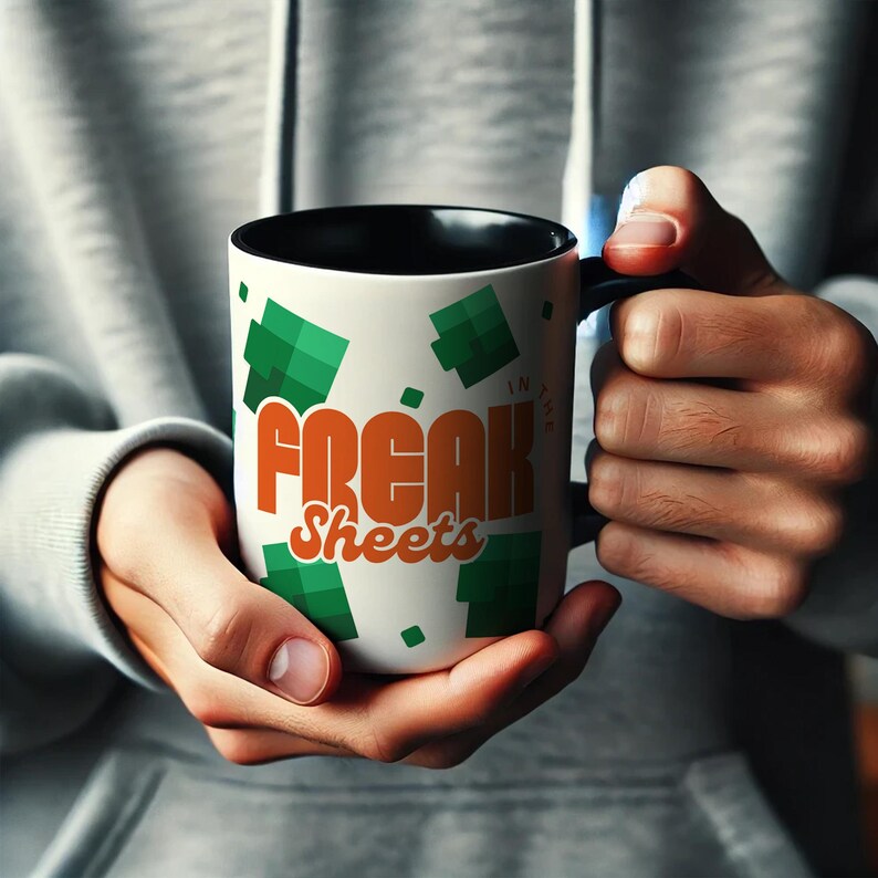 May include: A white coffee mug with a black rim and handle. The mug has a green and orange design with the text "Freak Sheets" and "In the" in a smaller font.