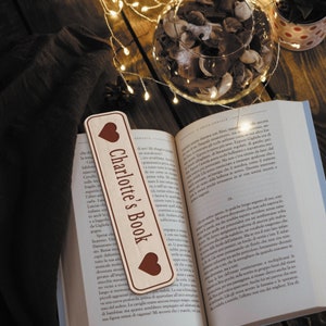 Op de afbeelding: Een houten bladwijzer met een hartvormig ontwerp en de tekst "Charlotte's Book" in bruine letters. De bladwijzer ligt op een boek met een bruine kaft.