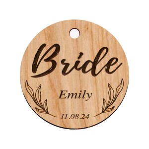 Puede incluir: Etiqueta redonda de madera con la palabra "Bride" en escritura cursiva, el nombre "Emily" y la fecha "11.08.24" grabados en ella.