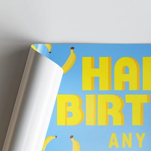 May include: A blue poster with yellow text that reads "HAPPY BIRTHDAY ANY". The poster features yellow banana illustrations. The corner of the poster is peeled back, revealing a white surface.