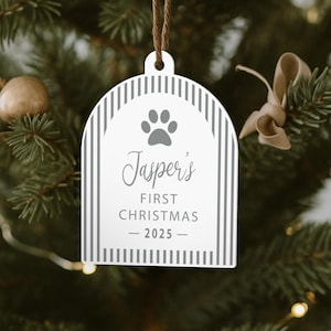 May include: A white Christmas ornament with a paw print and the text "Jasper's FIRST CHRISTMAS - 2025". The ornament has a striped border and is hanging from a brown string. The background is a blurred Christmas tree.