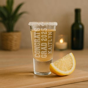 May include: A clear shot glass with a salted rim, filled with a golden liquid, and the text "CONGRATS GRAD 2025 KAITLYN" in white. A lemon wedge sits beside the glass on a wooden surface. A green bottle and a candle are in the background.
