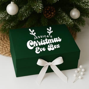 May include: A dark green rectangular gift box with a white bow and the text "JESSICA'S Christmas Eve Box". The box is decorated with white antlers and is placed in front of a Christmas tree with ornaments.