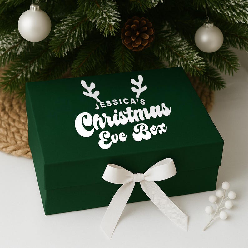 May include: A dark green rectangular gift box with a white bow and the text "JESSICA'S Christmas Eve Box". The box is decorated with white antlers and is placed in front of a Christmas tree with ornaments.