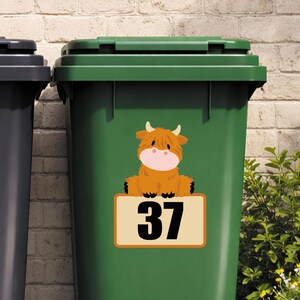 May include: A green trash bin with a cartoon Highland cow sticker. The sticker features a brown, fluffy cow with a pink snout and small horns, sitting above a rectangular sign with the number 37 in black.
