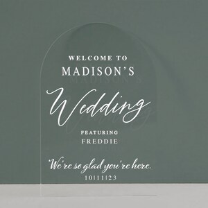 K&ouml;nnte beinhalten: Ein klares Acryl-Bogenschild mit dem Text "Willkommen zur Hochzeit von Madison mit Freddie" und "Wir freuen uns, dass Sie da sind." mit dem Datum 10.11.23.