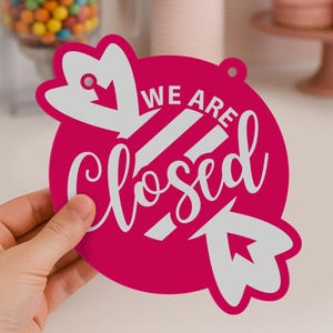 May include: A magenta-colored sign with the words "WE ARE Closed" in white script. The sign has a circular shape with decorative arrow-shaped elements and a small hole for hanging.