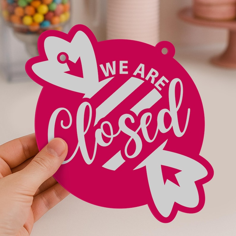 May include: A magenta-colored sign with the words "WE ARE Closed" in white script. The sign has a circular shape with decorative arrow-shaped elements and a small hole for hanging.