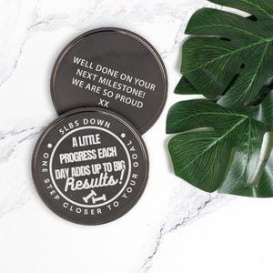 Puede incluir: Dos posavasos redondos negros con texto blanco. El posavasos superior dice "Well done on your next milestone! We are so proud XX". El posavasos inferior dice "5lbs down. A little progress each day adds up to big results! One step closer to your goal."