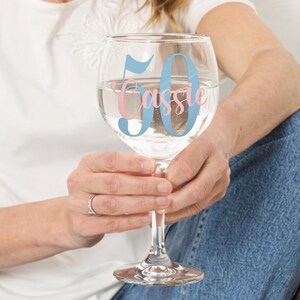May include: A clear, stemmed wine glass filled with liquid, featuring the number "50" in light blue and the name "Cassie" in pink. The glass is held by a person wearing a white shirt and blue jeans.