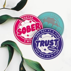 May include: Three round, colorful coasters with motivational messages. The pink coaster says "SOBER 6 MONTHS ONE DAY AT A TIME". The blue coaster says "TRUST THE PROCESS ONE DAY AT A TIME". The turquoise coaster says "1 YEAR SOBER".