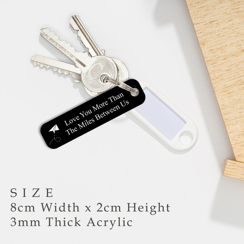 Puede incluir: Llavero de acr&iacute;lico negro con el texto "Love You More Than The Miles Between Us" y un peque&ntilde;o gr&aacute;fico de avi&oacute;n de papel. El llavero mide 8 cm de ancho por 2 cm de alto y tiene un grosor de 3 mm.