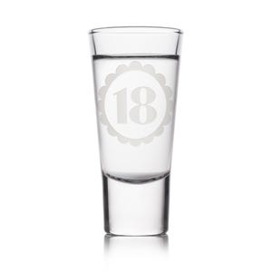 May include: A clear shot glass with the number 18 etched in a scalloped circle. The glass is filled with a clear liquid. The shot glass is a perfect gift for an 18th birthday celebration.