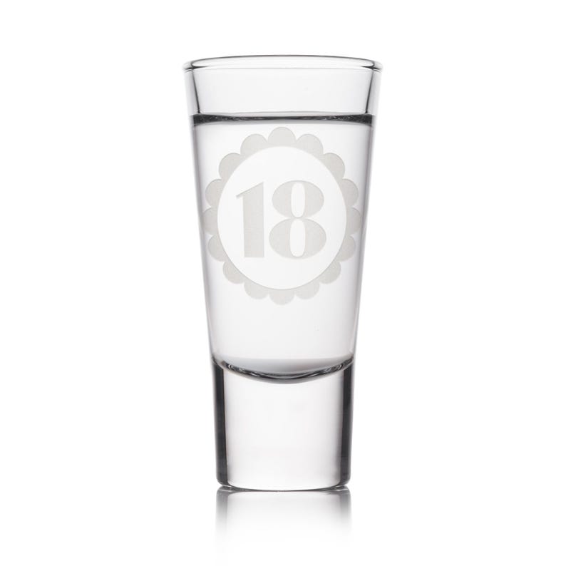 May include: A clear shot glass with the number 18 etched in a scalloped circle. The glass is filled with a clear liquid. The shot glass is a perfect gift for an 18th birthday celebration.