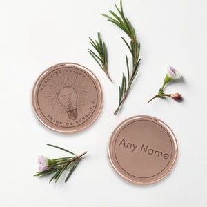 Puede incluir: Dos posavasos redondos de color oro rosa. Un posavasos tiene el texto "TEACHING BRIGHT STUDENTS TO SHINE" alrededor de un gr&aacute;fico de bombilla. El otro posavasos tiene el texto "Any Name". Ramitas verdes y peque&ntilde;as flores est&aacute;n esparcidas alrededor de los posavasos.