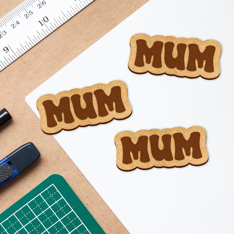 May include: Three brown wooden cutout signs with the word "MUM" in a retro font. The signs are on a white surface.