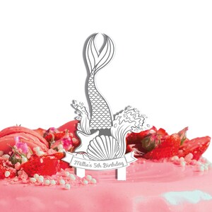 May include: White cake topper with a mermaid tail, a seashell, and waves. The topper has a banner that says "Millie's 5th Birthday".