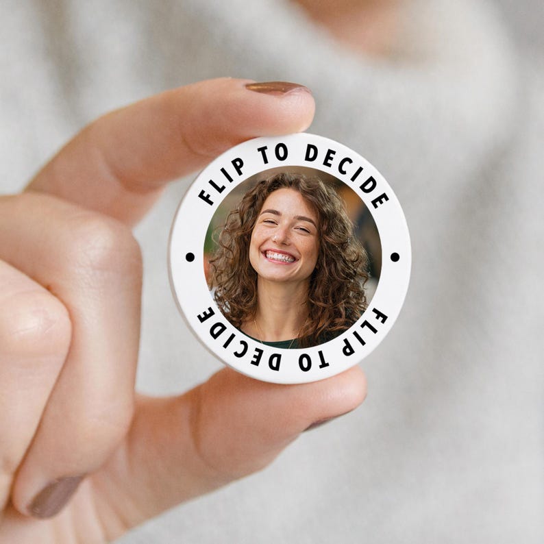 May include: A white circular button with the text "FLIP TO DECIDE" encircling a smiling woman's photo. Two black dots are on either side of the photo. The button is held by a person.