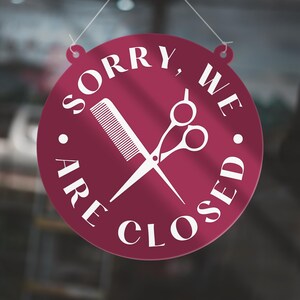 May include: A round, burgundy sign with white text and a white comb and scissors graphic. The sign reads "SORRY, WE ARE CLOSED" in a circular arrangement, indicating a closed business, likely a salon or barber shop.