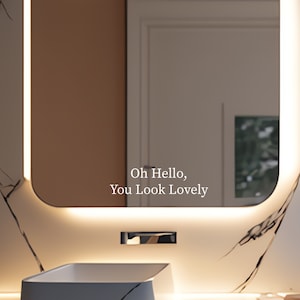 Peut inclure: Une vasque de salle de bain moderne avec un lavabo blanc et un grand miroir &eacute;clair&eacute; avec un bord arrondi. Le miroir porte l'inscription "Oh Hello, You Look Lovely".