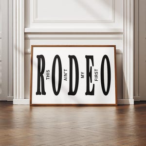 May include: Black and white typography print with the word "RODEO" and the text "This ain't my first" written vertically within the letters.