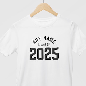 May include: White t-shirt with the text "ANY NAME CLASS OF 2025" in black. The text is centered on the front of the shirt. The shirt is hanging on a wooden hanger.