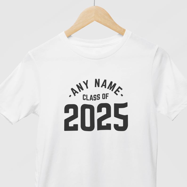 May include: White t-shirt with the text "ANY NAME CLASS OF 2025" in black. The text is centered on the front of the shirt. The shirt is hanging on a wooden hanger.