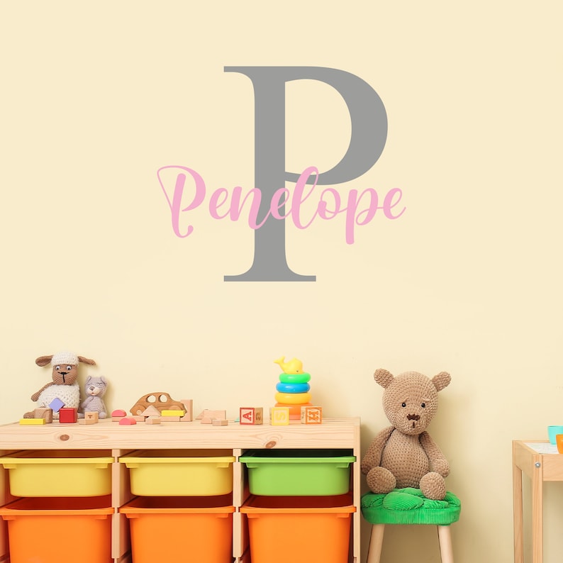 May include: A gray letter P with the name Penelope written in pink cursive underneath. The wall is a light yellow color. There are toys on a wooden shelf below the wall decal.