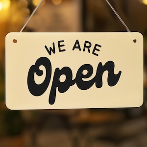 May include: A rectangular cream-colored sign with the words "WE ARE" above and "Open" in large, bold, black letters. The sign is hanging by two thin strings, with a blurred background.