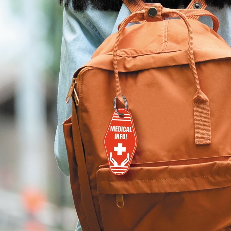 Pode incluir: Uma mochila marrom com um chaveiro vermelho que diz "MEDICAL INFO!" com uma cruz branca e m&atilde;os ao redor.