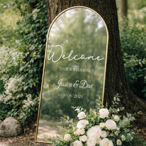 May include: A large, arched mirror with a gold frame displays the words "Welcome to our Wedding" and the names "Jessica & Dan" along with the date "10/08/2026". The mirror is surrounded by greenery and white roses.