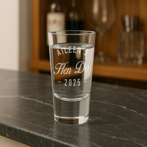 May include: A clear shot glass with the text "Aileen's Hen Do - 2025" etched on the front. The glass is filled with a clear liquid and sits on a dark countertop. The background includes a wooden cabinet and other glassware.