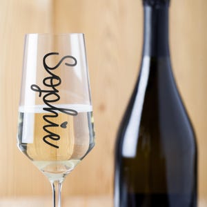 May include: A clear glass champagne flute with the name "Sophie" written on the side in black cursive script. The flute is filled with a sparkling white beverage. A bottle of champagne is in the background.