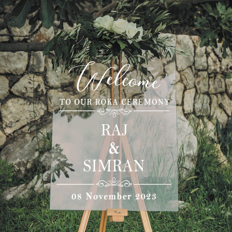 May include: A clear acrylic welcome sign with white text. The sign reads "Welcome to our Roka Ceremony Raj & Simran 08 November 2023".