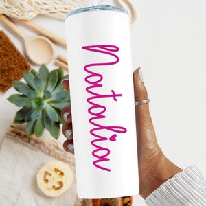 May include: White stainless steel tumbler with a pink inscription that reads "Natalia" with a small heart symbol.