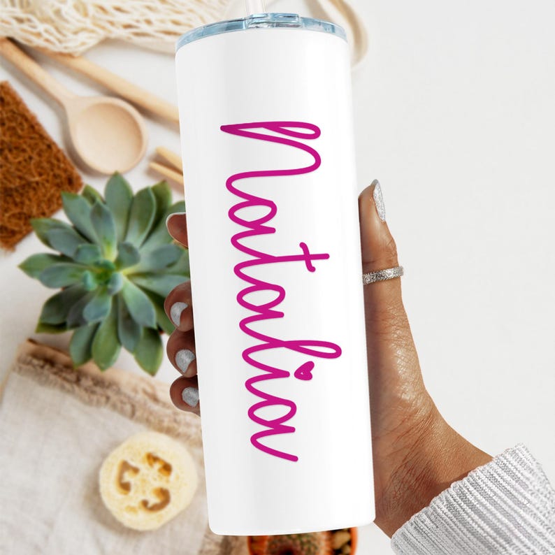 May include: White stainless steel tumbler with a pink inscription that reads "Natalia" with a small heart symbol.