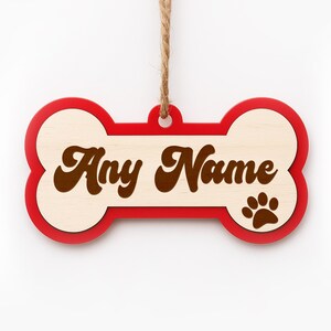 Pode incluir: Enfeite de madeira em forma de osso de c&atilde;o com o texto "Any Name" em escrita castanha. O osso tem uma borda vermelha e uma pegada na pata direita. Uma argola de barbante &eacute; presa para pendurar.