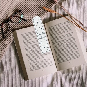 May include: A white bookmark with a rounded top and a black outline. The bookmark has the text "book babe" in black lettering and a star design. The bookmark is placed on a book with a white background.