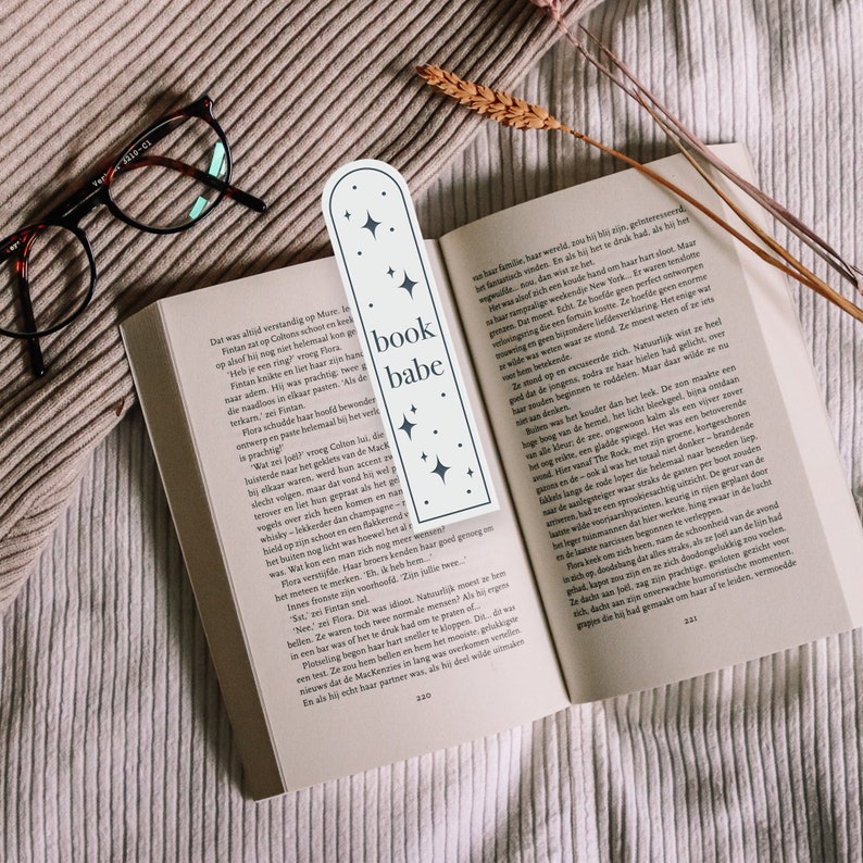 May include: A white bookmark with a rounded top and a black outline. The bookmark has the text "book babe" in black lettering and a star design. The bookmark is placed on a book with a white background.