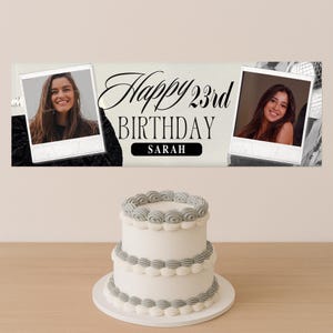Op de afbeelding: Een witte verjaardagstaart met twee lagen en grijze glazuuraccenten staat op een houten tafel. Boven de taart staat een banner met de tekst "Happy 23rd Birthday Sarah" en twee ingelijste foto's van een vrouw.