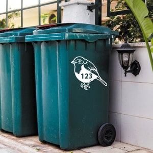 May include: Dark teal trash bin with a white bird decal and the number 123. The bin has a black wheel and a dark green lid. A second bin is partially visible. A black outdoor lamp is mounted on the wall.