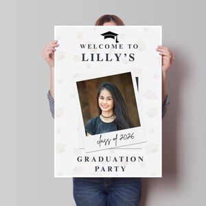 May include: A graduation party sign held by a person. The sign is white with the text "WELCOME TO LILLY'S" and "GRADUATION PARTY". A photo of a smiling person is in the center, with the text "class of 2026" below it.
